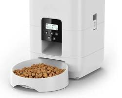 A modern white automatic pet feeder dispensing dry food kibble into a stainless steel bowl on a clean interior floor.